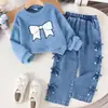 Estilo europeu e americano: um conjunto de roupa doce e delicado para uma menina, apresentando um top branco, macio e de manga comprida com um design de laço lateral completo, combinado com calças jeans - um conjunto de duas peças