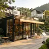 Cafetería tipo contenedor de estilo industrial con muros cortina de cristal de suelo a techo y una plataforma de observación en el nivel superior, ubicada en un distrito cultural y comercial