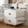 Mesita de noche de color crema con acolchado suave, esquinas redondeadas, doble cajón y patas de madera maciza, un mueble de almacenamiento simple y práctico para el dormitorio