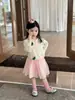 Coleção Outono 2026: Roupas infantis estilo coreano, apresentando um cardigã de tricô com desenho animado e uma saia fofa de tule com pompons para meninas