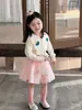 Coleção Outono 2026: Roupas infantis estilo coreano, apresentando um cardigã de tricô com desenho animado e uma saia fofa de tule com pompons para meninas