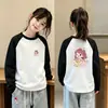 Camiseta infantil moderna e estilosa com blocos de cor, gola redonda, mangas compridas e estampa de desenho animado, perfeita como camada base para o outono e inverno de 2026