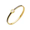 Este elegante brazalete presenta un refinado acabado dorado con una llamativa franja de esmalte azul y un centro incrustado con una reluciente gema blanca Su diseño atemporal asegura que realza cualquier atuendo añadiendo un toque de color y sofisticación Perfecto para el uso diario ocasiones especiales o como un regalo considerado