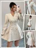 Vestido Chanel 2026 na cor aveia, decote em V, botões de pérola duplos, mangas compridas, acinturado e com barra larga, estilo socialite francesa