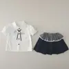 Nuevo atuendo de verano 2026 para niñas: conjunto elegante de manga corta con cuello, que presenta una camisa de dibujos animados y una falda corta, disponible para venta al por mayor